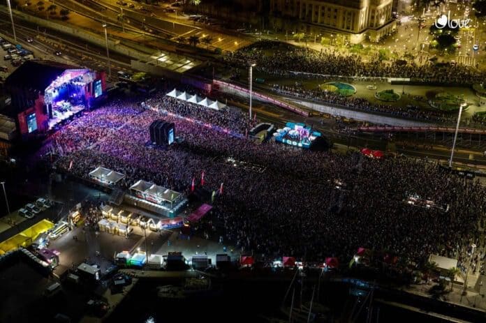 Vista aèria i panoràmica nocturna d'un festival de música multitudinari a la ciutat, amb un gran escenari il·luminat a la part esquerra i una immensa multitud que omple tot el recinte.