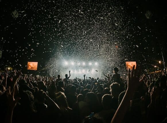 Una gran multitud en un concert nocturn a l'aire lliure, vista des del darrere, amb els braços aixecats mentre una pluja massiva de confeti blanc cau sota els llums brillants de l'escenari.
